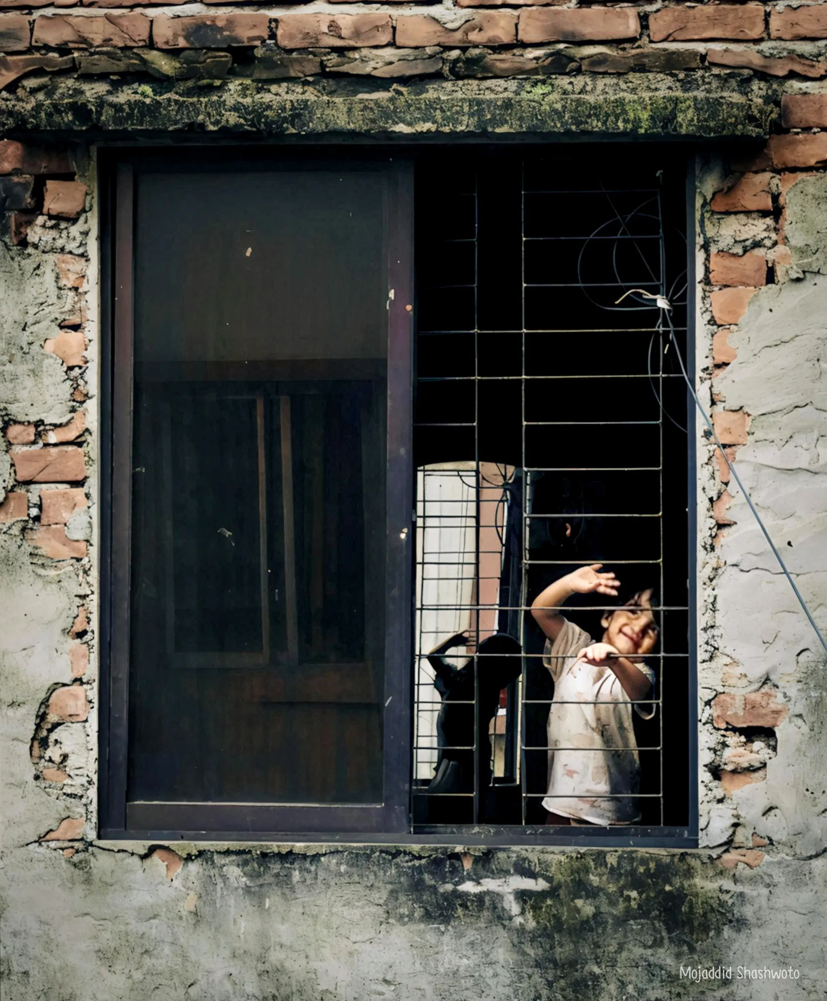 Child waving from behind a dark window grille in a rustic brick wall