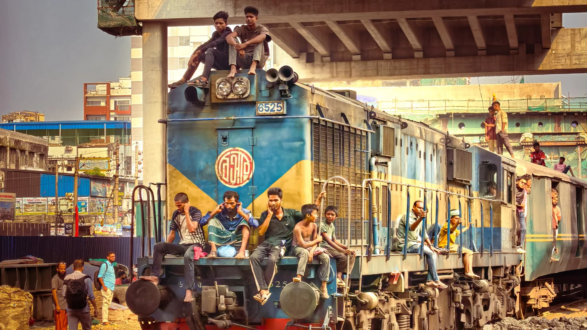 Passengers and commuters riding on a blue diesel locomotive under an elevated flyover in Dhaka