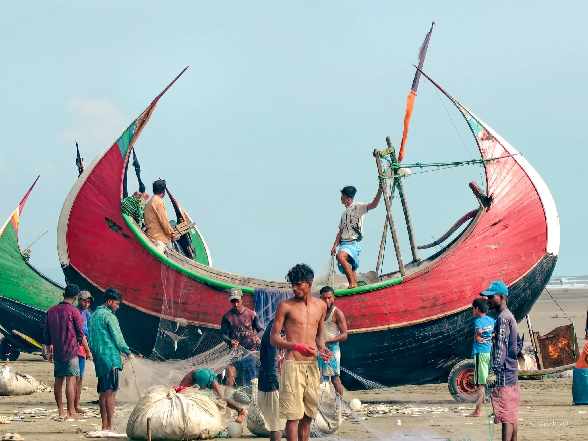 Fishermen hauling white nets beside a large red crescent moon boat on the shore